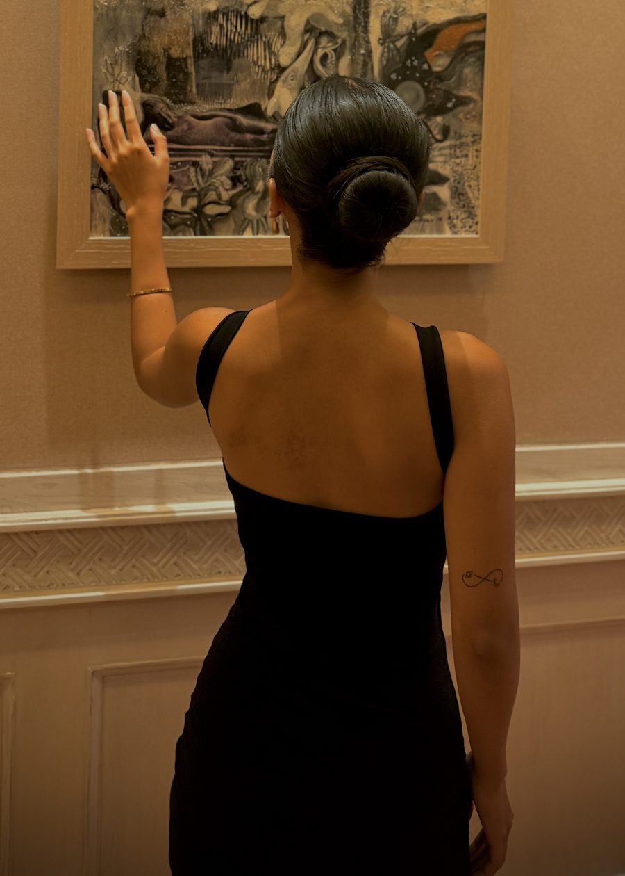 Fera in an open-back black Ophelia dress, reaching toward a painting in a gallery